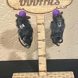 May include: A pair of black rat earrings with purple hats, hanging from a wooden sign that reads "Demelo's Oddities".