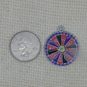 May include: A silver charm with a colorful wheel of fortune design. The wheel has red, black, purple, and yellow sections with various symbols. The charm is about the size of a US quarter.