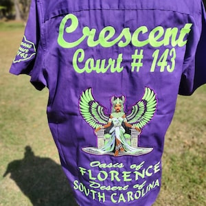 May include: A purple short-sleeved button-down shirt featuring bright green embroidery. The back displays the text Crescent Court # 143, an illustration of a winged figure on a throne, and the text Oasis of FLORENCE Desert of SOUTH CAROLINA.