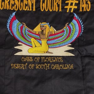 May include: Black embroidered shirt with a gold design of a woman with wings, a pyramid, and the text "Crescent Court #143 Oasis of Florence Desert of South Carolina".