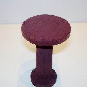 May include: A small, round, wooden table with a purple painted finish. The table has a single, thick, cylindrical leg.
