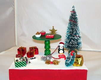 May include: A miniature Christmas scene with a green table, red and green presents, a small penguin figurine, a frosted Christmas tree, and gold tinsel.