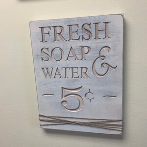 May include: White wooden sign with the text "FRESH SOAP & WATER - 5&cent; -" in a vintage style font. The sign is decorated with twine.