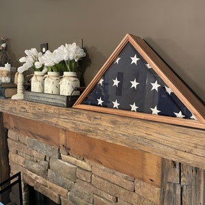 May include: A wooden mantelpiece with a framed American flag, white flowers in jars, and a decorative candle on top. The mantel is above a stone fireplace.