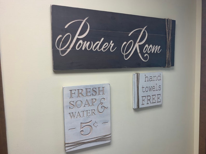 May include: Three wooden signs with white lettering on a beige wall. The top sign reads "Powder Room". The middle sign reads "Fresh Soap & Water - 5&cent;". The bottom sign reads "hand towels FREE".