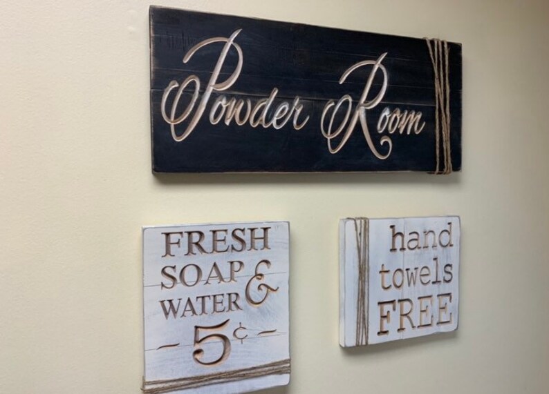 May include: Three rustic wooden signs with white lettering. The top sign reads "Powder Room" and is black. The middle sign reads "FRESH SOAP & WATER - 5&cent; -" and is white. The bottom sign reads "hand towels FREE" and is white.