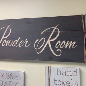 May include: A dark gray wooden sign with the words "Powder Room" in white lettering. The sign is attached to the wall with twine.