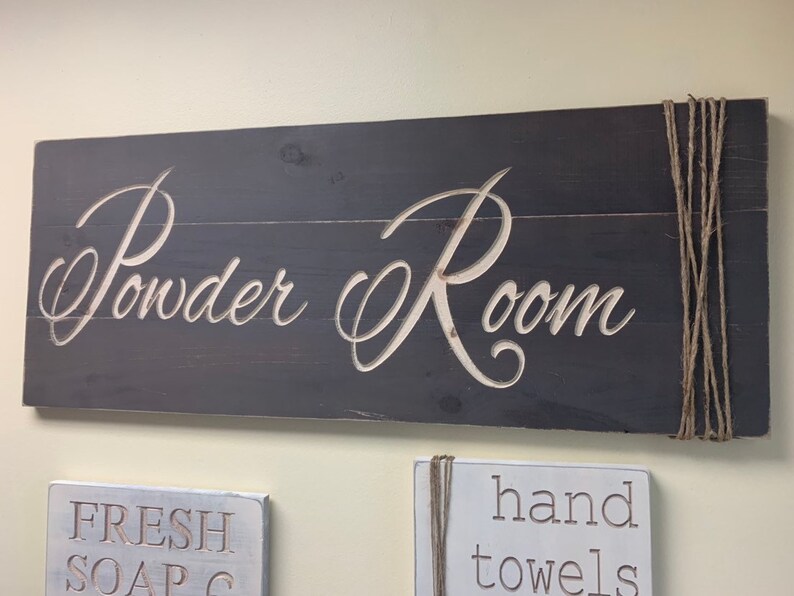 May include: A dark gray wooden sign with the words "Powder Room" in white lettering. The sign is attached to the wall with twine.