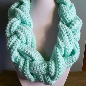 May include: A mint green, chunky knit infinity scarf displayed on a white mannequin head. The scarf is made of soft yarn and features a braided design. The handmade accessory is perfect for adding warmth and style to any outfit.