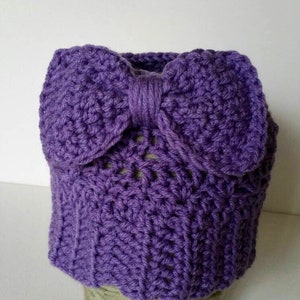May include: A handmade, purple crochet hat with a large bow detail. The hat is made of a textured yarn and sits atop a ball of green yarn. The bow is centered on the front of the hat.