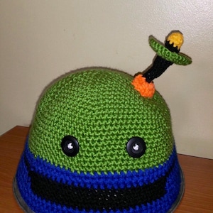 May include: Green crocheted hat with black and blue stripes, two black buttons for eyes, and a yellow and orange crocheted antenna on top.