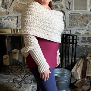 May include: A beige, crocheted shawl with long sleeves. The shawl is worn over a burgundy top and blue pants. The background features a stone fireplace, a metal bucket, and firewood.
