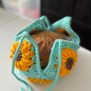 May include: A crocheted bread basket with a teal blue base and yellow sunflower accents. The basket is holding a loaf of bread.