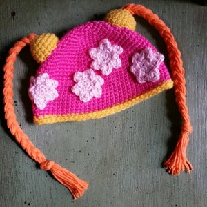May include: A handmade crochet hat in bright pink, featuring three light pink flower appliqués. The hat has a yellow brim and two yellow pom-poms. Long orange braids with tassels extend from the hat, creating a playful and whimsical design.