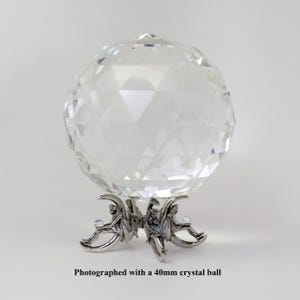 Puede incluir: Una bola de cristal transparente y facetada, de aproximadamente 4 cm de di&aacute;metro, se encuentra sobre un soporte decorativo de metal plateado. El soporte presenta tres figuras. El texto en la parte inferior dice: Photographed with a 40mm crystal ball.