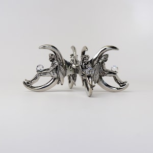 Puede incluir: Pinza para el pelo de metal plateado con tres figuras aladas, cada una sosteniendo un cristal transparente. Las figuras est&aacute;n sentadas sobre formas de media luna. La pinza tiene un dise&ntilde;o detallado y ornamentado.