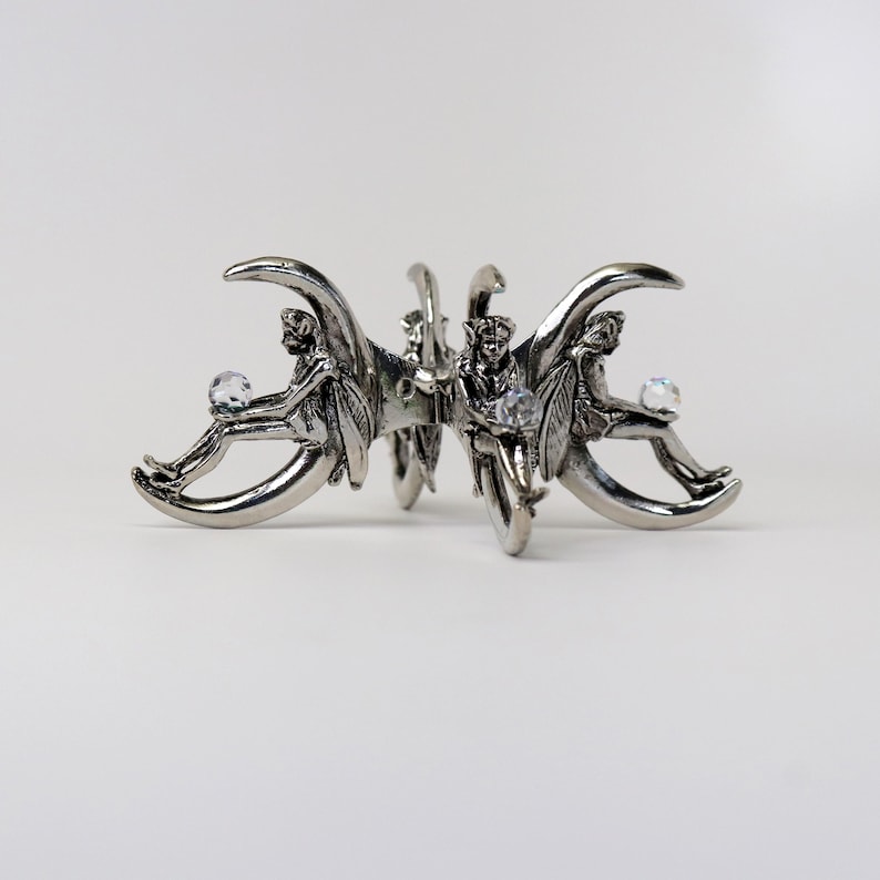 Puede incluir: Pinza para el pelo de metal plateado con tres figuras aladas, cada una sosteniendo un cristal transparente. Las figuras est&aacute;n sentadas sobre formas de media luna. La pinza tiene un dise&ntilde;o detallado y ornamentado.