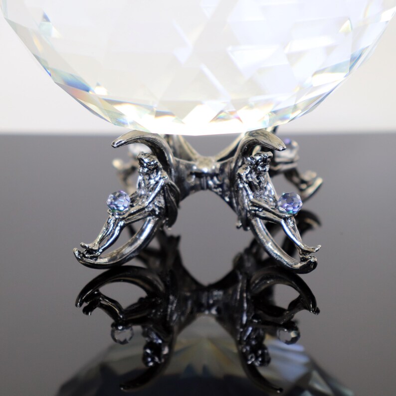 Puede incluir: Una bola de cristal clara y facetada descansa sobre un soporte de metal plateado. El soporte presenta cuatro figuras, cada una sosteniendo un peque&ntilde;o cristal azul claro. La superficie reflectante de abajo refleja el dise&ntilde;o. Art&iacute;culo decorativo.