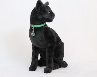 May include: A black plush cat toy wearing a green collar with a shamrock charm. The cat is sitting with its head turned to the side.