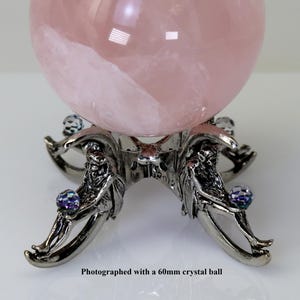 Puede incluir: Una gran bola de cristal rosa transl&uacute;cido descansa sobre un soporte decorativo de color plateado. El soporte presenta intrincados detalles de figuras que sostienen peque&ntilde;as esferas de cristal iridiscentes. El texto en la parte inferior dice: Fotografiado con una bola de cristal de 60 mm.