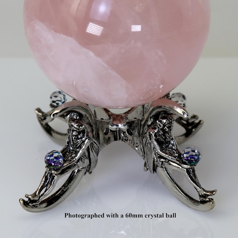 Puede incluir: Una gran bola de cristal rosa transl&uacute;cido descansa sobre un soporte decorativo de color plateado. El soporte presenta intrincados detalles de figuras que sostienen peque&ntilde;as esferas de cristal iridiscentes. El texto en la parte inferior dice: Fotografiado con una bola de cristal de 60 mm.