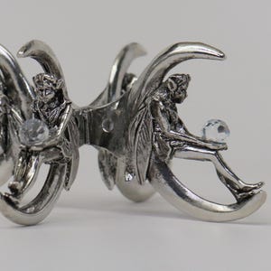 Puede incluir: Una pinza para el pelo plateada con figuras de hadas sentadas en forma de media luna. Cada hada sostiene una bola de cristal clara y facetada. La pinza tiene un dise&ntilde;o detallado y ornamentado.