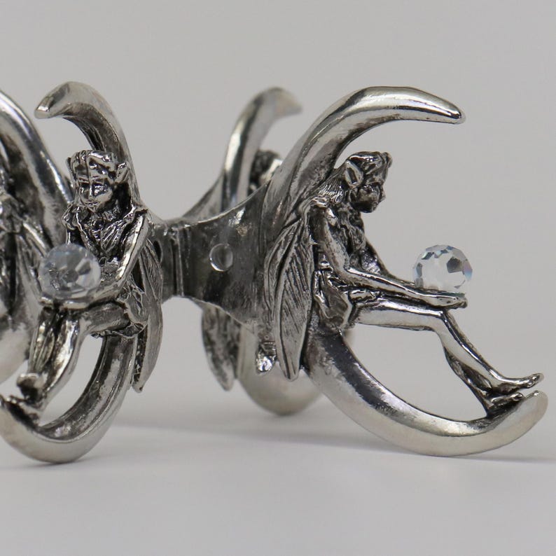 Puede incluir: Una pinza para el pelo plateada con figuras de hadas sentadas en forma de media luna. Cada hada sostiene una bola de cristal clara y facetada. La pinza tiene un dise&ntilde;o detallado y ornamentado.