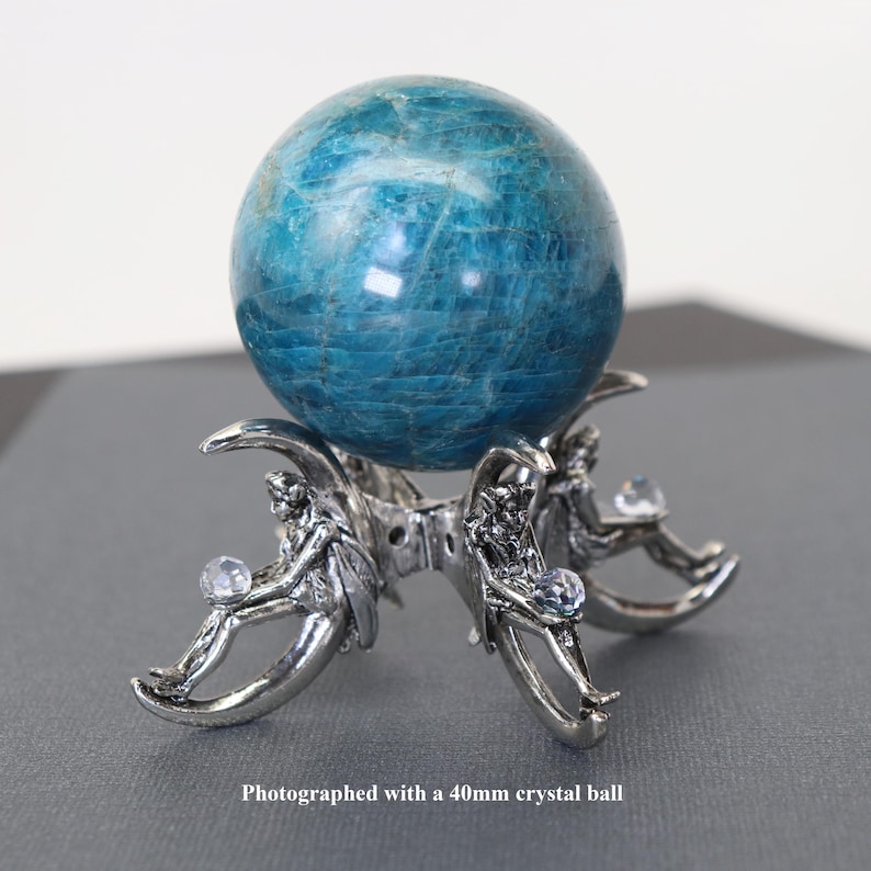 Puede incluir: Una bola de cristal turquesa, de aproximadamente 4 cm de di&aacute;metro, descansa sobre un soporte plateado. El soporte presenta tres figuras y detalles de cristal transparente. El texto en la parte inferior dice: "Photographed with a 40mm crystal ball."
