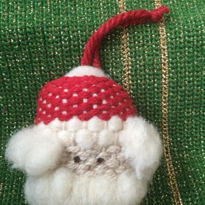 Puede incluir: Un adorno de Santa Claus de fieltro blanco con un gorro rojo y blanco y una barba blanca. El adorno tiene una cuerda roja para colgar.