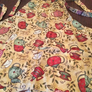 Könnte beinhalten: Eine beige Schürze mit einer Tasche und verstellbaren Trägern. Die Schürze ist mit einem sich wiederholenden Muster aus Teekannen und Teetassen mit floralen Akzenten verziert. Der Text auf der Schürze lautet "green tea", "peppermint", "sugar plum", "chamomile", "English Breakfast" und "green".