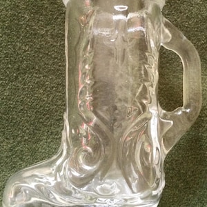 May include: Clear glass boot-shaped mug with a handle and a decorative floral pattern. The mug has a clear rim and is designed to resemble a boot. The glass is transparent, allowing the contents to be visible.