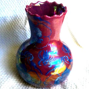 Peut inclure: Un vase d&eacute;coratif avec un bord &eacute;vas&eacute; et festonn&eacute;. Le vase a une couleur de base rouge fonc&eacute; avec des motifs floraux bleus et des reflets iris&eacute;s. Le vase a un corps bulbeux et un col fin.