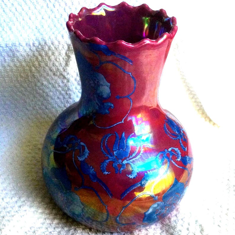 Peut inclure: Un vase d&eacute;coratif avec un bord &eacute;vas&eacute; et festonn&eacute;. Le vase a une couleur de base rouge fonc&eacute; avec des motifs floraux bleus et des reflets iris&eacute;s. Le vase a un corps bulbeux et un col fin.