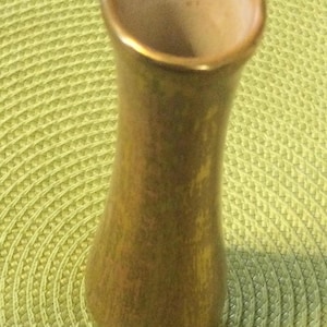 May include: A small, decorative vase with a flared top and a slender body. The vase has a gold-colored rim and a muted, earthy tone, with a subtle gradient from a darker base to a lighter top. The vase is likely ceramic or glass.