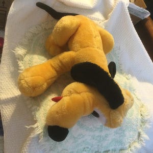 Puede incluir: Un peluche de Pluto, un perro amarillo con orejas y cola negras, y una lengua roja. El juguete está sobre una superficie blanca y azul claro.