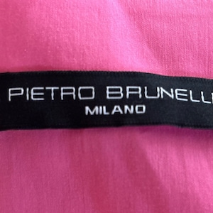 以下が含まれることがあります： 白い文字で「PIETRO BRUNELLI MILANO」と書かれた黒いラベル。ラベルはピンク色の生地の背景に配置されています。生地は滑らかで織られた素材のようです。