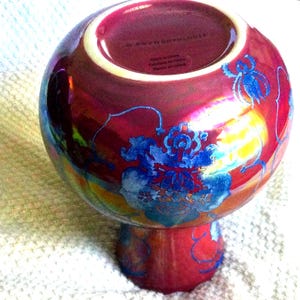 Peut inclure: Un vase en c&eacute;ramique rouge iris&eacute; avec un dessus rond et une base en pi&eacute;destal. Le vase est d&eacute;cor&eacute; de motifs floraux bleus. Le bas du vase porte l'inscription "ANTHROPOLOGIE" et "Made in China".