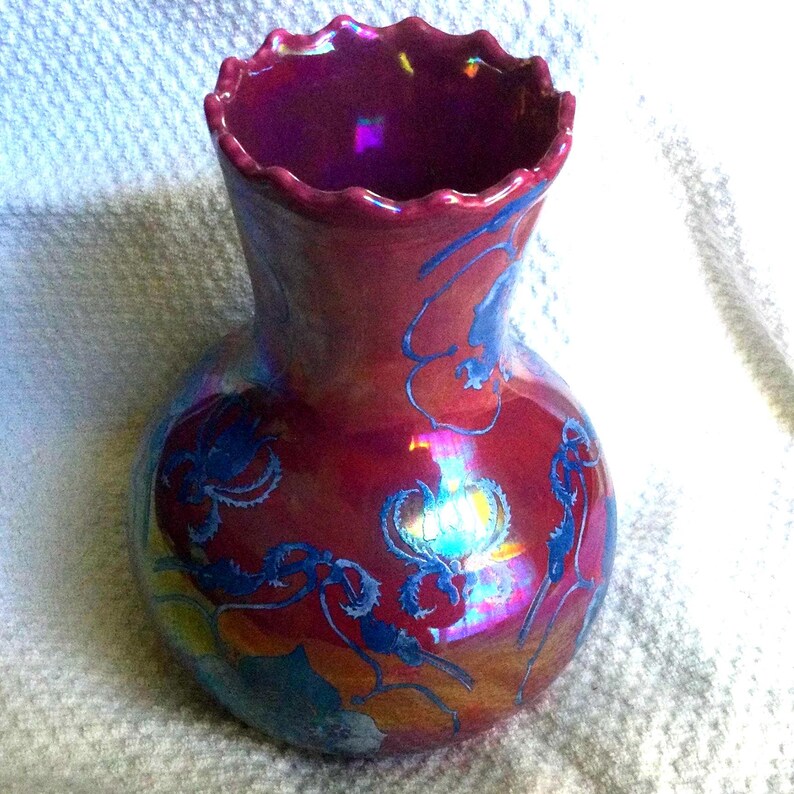 Peut inclure: Un vase d&eacute;coratif avec un corps bulbeux et un bord festonn&eacute;. Le vase est d'un rouge profond avec des reflets iris&eacute;s et des motifs floraux bleus. Le vase est probablement en c&eacute;ramique ou en verre.