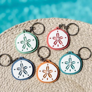May include: A collection of colorful sand dollar keychains. Each keychain features a white sand dollar design with a colored border in shades of blue, green, orange, and pink. The keychains are attached to silver key rings.