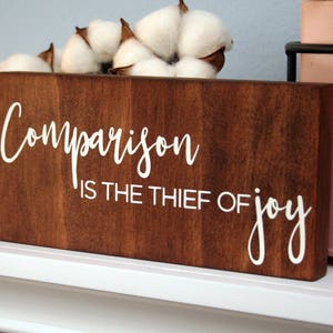 Puede incluir: Un letrero de madera con el texto "Comparison is the thief of joy" en letras blancas. El letrero es marr&oacute;n y tiene un aspecto r&uacute;stico.