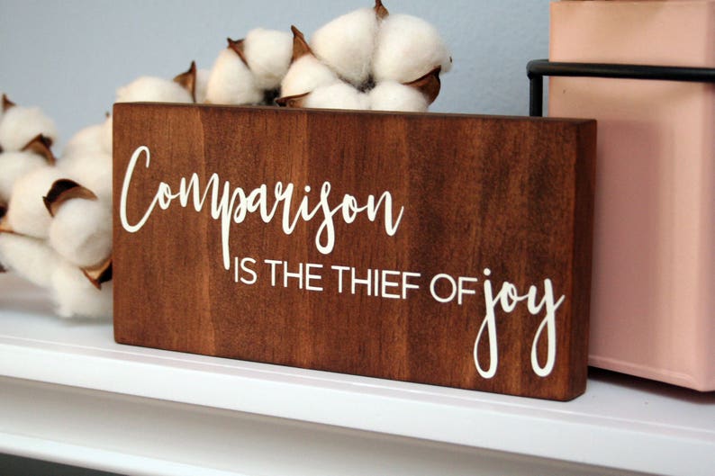 Puede incluir: Un letrero de madera con el texto "Comparison is the thief of joy" en letras blancas. El letrero es marr&oacute;n y tiene un aspecto r&uacute;stico.