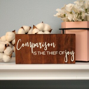 Puede incluir: Un letrero de madera con el texto "Comparison is the thief of joy" en letras blancas. El letrero es marr&oacute;n y tiene un aspecto r&uacute;stico. El letrero est&aacute; colocado en un estante blanco con bolas de algod&oacute;n y una maceta rosa con flores blancas.