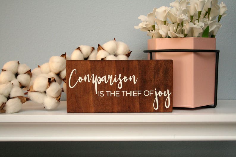 Puede incluir: Un letrero de madera con el texto "Comparison is the thief of joy" en letras blancas. El letrero es marr&oacute;n y tiene un aspecto r&uacute;stico. El letrero est&aacute; colocado en un estante blanco con bolas de algod&oacute;n y una maceta rosa con flores blancas.
