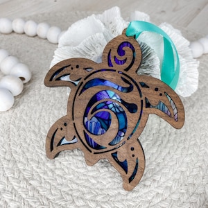 May include: A laser-cut wooden sea turtle ornament featuring a dark brown wood frame with intricate cutouts. The interior displays a vibrant, multi-colored stained glass-style insert in shades of blue, purple, and teal, finished with a teal ribbon.