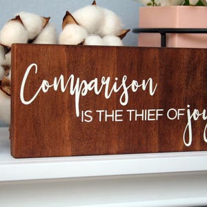 Puede incluir: Un letrero de madera con el texto "Comparison is the thief of joy" en letras blancas sobre un fondo marr&oacute;n.
