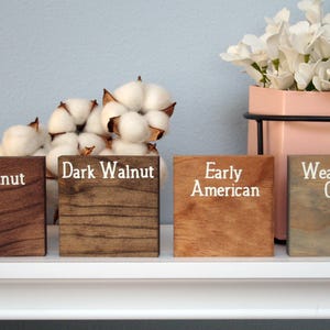 Puede incluir: Cuatro bloques de madera te&ntilde;idos en diferentes colores: Casta&ntilde;o, Nogal oscuro, Americano antiguo y Gris desgastado. Los bloques est&aacute;n dispuestos en una fila sobre un estante blanco.