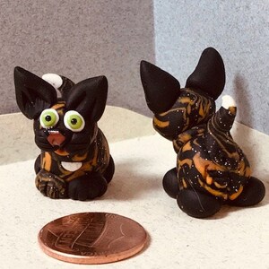 May include: Two small black and orange cat figurines with green eyes. The cats are made of polymer clay and have a speckled finish.