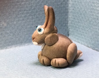 May include: A small, gray, clay rabbit figurine with blue eyes. The rabbit is sitting with its ears upright and its tail tucked behind its legs.