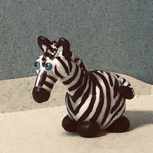 May include: A small, hand-crafted zebra figurine made of black and white clay with blue eyes. The zebra is sitting on a white surface.