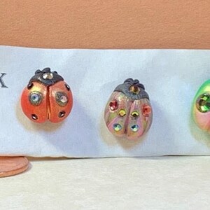 May include: Three ladybug-shaped pins with different colored bodies and jeweled accents. The pins are on a white card with the letter "K" printed on it.
